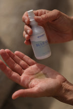 Load image into Gallery viewer, Dresden Holden Wellness Body + Welless SAnta Barbara based acupuncture and skincare studio. A blue face a body oil in the palm of a hand, the other hand is holding a 2 ounce white glass bottle with a green and blue label that says Blue Oil

